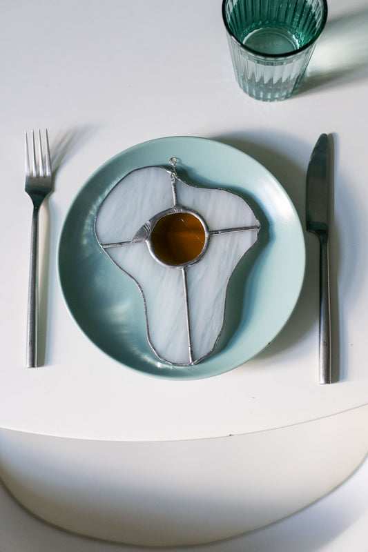 A stained glass art work in the shape of a sunny side up egg on a blue ceramic plate on a whitetable with a glass and cutlery.