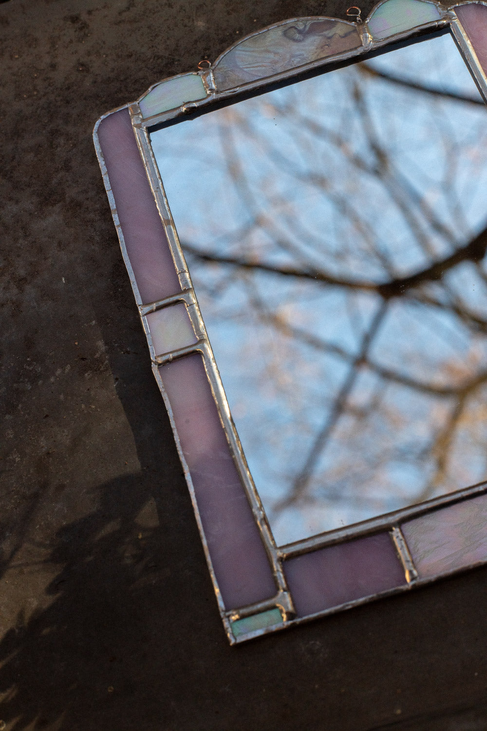 Stained glass mirror with a pink frame reflecting tree branches against a blue sky.