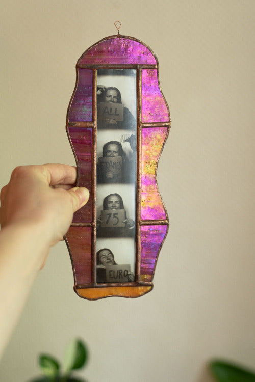 Hand holding a colorful stained glass frame with cut-out portraits against a beige wall, held by a hand.