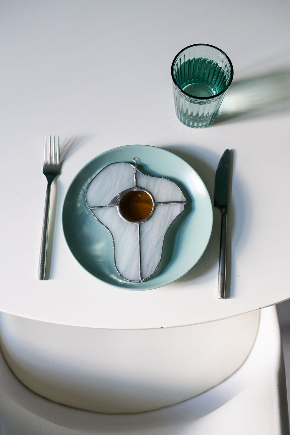 A stained glass art work in the shape of a sunny side up egg on a blue ceramic plate on a whitetable with a glass and cutlery.