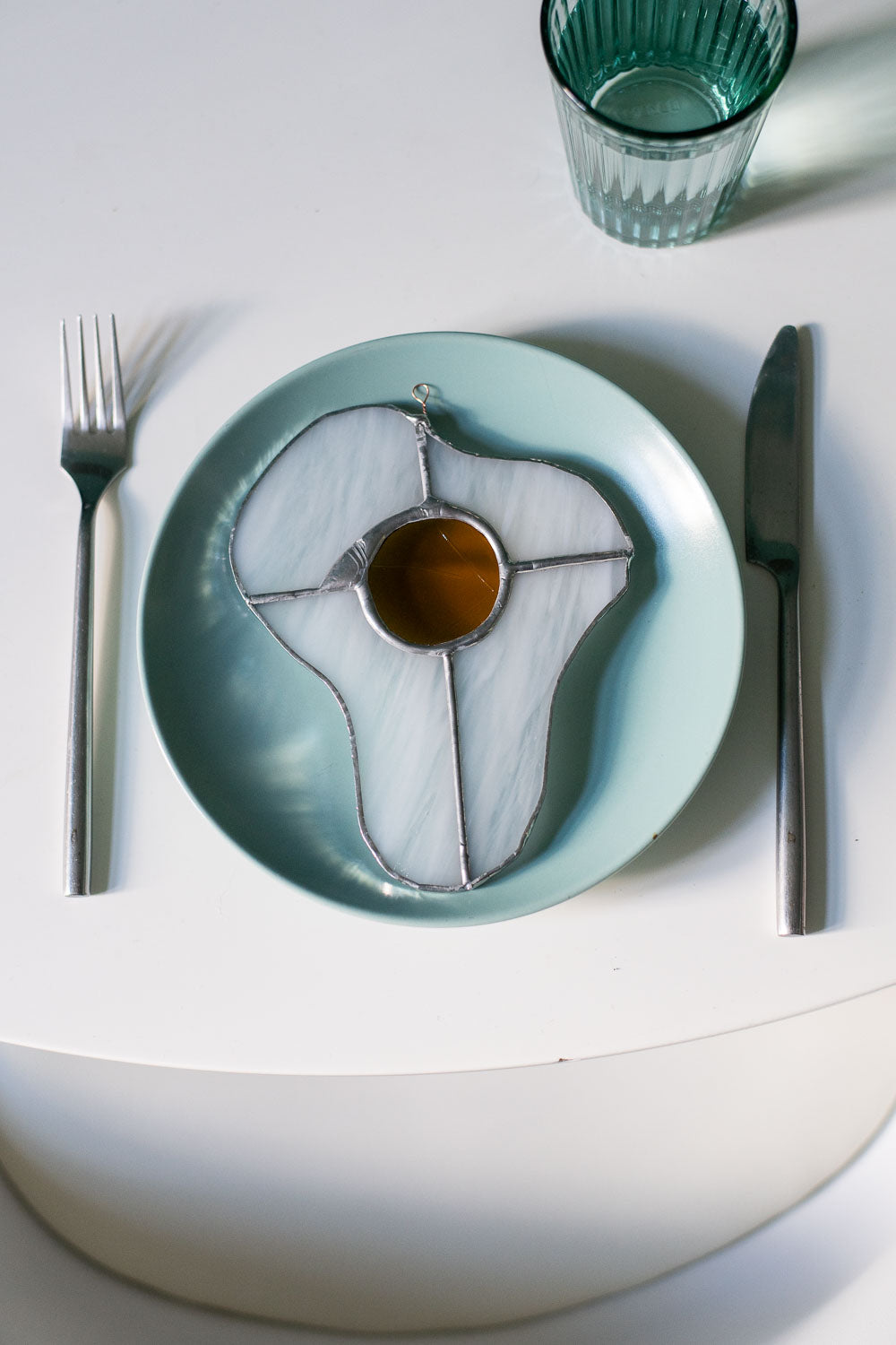 A stained glass art work in the shape of a sunny side up egg on a blue ceramic plate on a whitetable with a glass and cutlery.