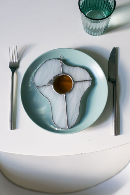 A stained glass art work in the shape of a sunny side up egg on a blue ceramic plate on a whitetable with a glass and cutlery.