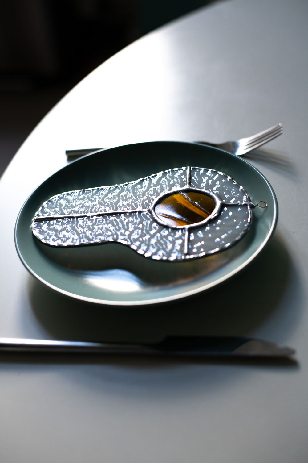 Teal plate with a decorative egg-shaped stained glass artwork, fork, knife, and glass on a white surface.