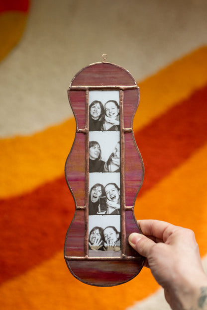 Colorful stained glass frame with photobooth photos inside, held by a hand against a beige background