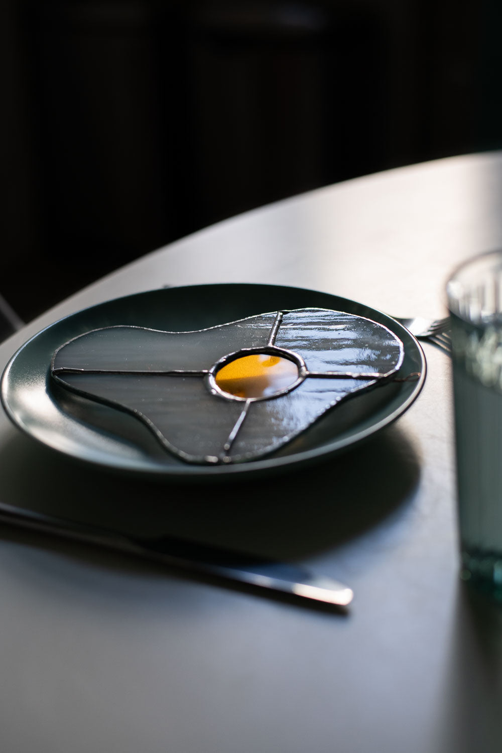 Teal plate with a decorative egg-shaped stained glass artwork, fork, knife, and glass on a white surface.