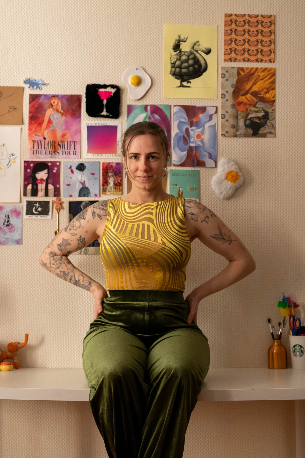 Woman in a yellow top and green pants standing in front of a wall with colorful posters.