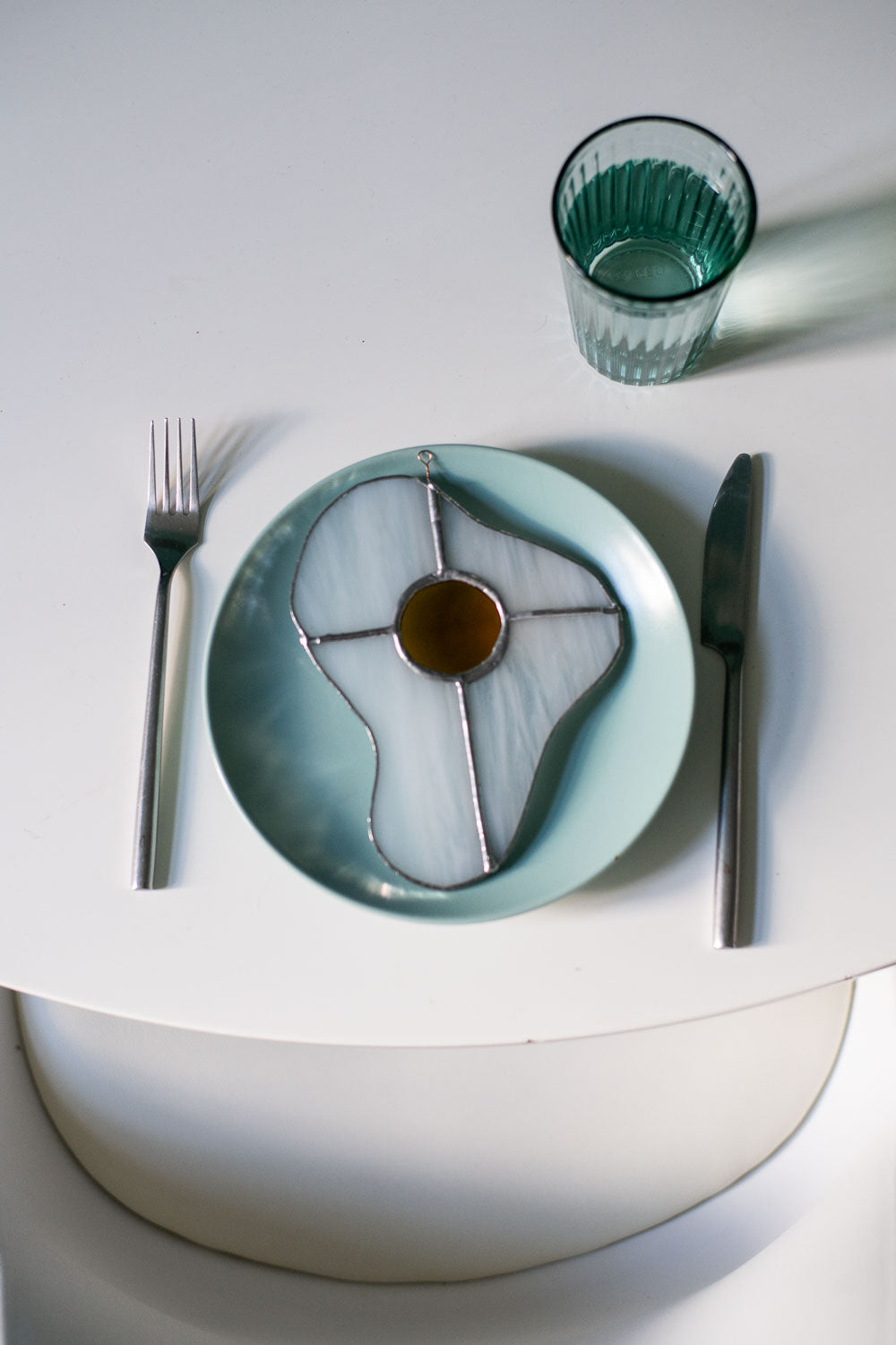Teal plate with a decorative egg-shaped stained glass artwork, fork, knife, and glass on a white surface.